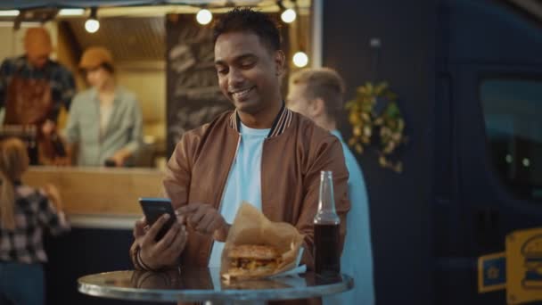 Yakışıklı genç Hintli adam açık hava sokak yemekhanesinde bir masada otururken akıllı telefon kullanıyor. İnterneti ya da sosyal medyayı tarıyor, bir durum güncellemesi yayınlıyor. İnsanlar mutlu ve gülümsüyor.. — Stok video