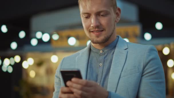 Bonito jovem homem concentrado está usando um smartphone enquanto está de pé ao ar livre na noite. Ele está navegando na Internet ou nas mídias sociais, postando uma atualização de status. O homem está vestindo um terno . — Vídeo de Stock