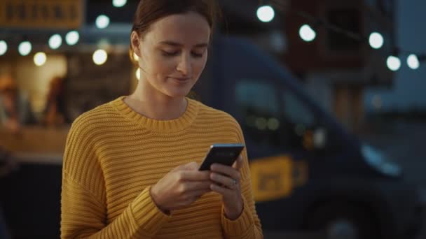 Hermosa morena joven mujer está utilizando un teléfono inteligente mientras que de pie al aire libre en la noche. Está navegando por Internet o las redes sociales, publicando una actualización de estado. Ella mira hacia otro lado mientras piensa . — Vídeo de stock