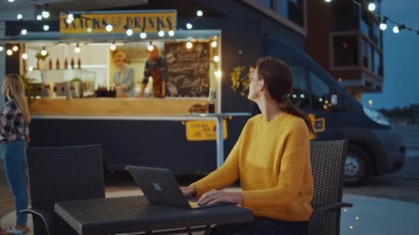 Belle jeune femme brune travaille sur ordinateur portable tout en s'asseyant à une table dans un café de nourriture de rue en plein air. Elle navigue sur Internet ou sur les réseaux sociaux, affichant une mise à jour de situation. Attendre son ordre . — Video