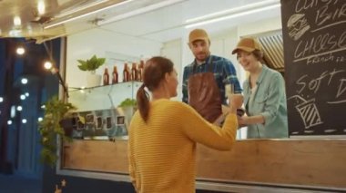 Yemek kamyonu çalışanı Mutlu Genç Hipster Müşterilerine Yeni Yapılmış Biftek Burgerleri, Kızartmalar ve Soğuk İçecekler Veriyor. Modern Havalı Mahallede Sokak Yemeği Satan Ticari Kamyon.