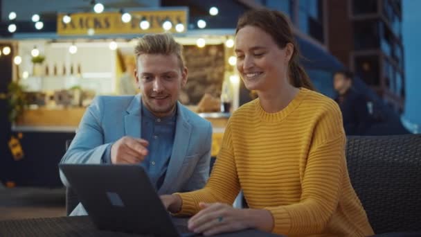 Vackra Unga Par använder en bärbar dator medan du sitter vid ett bord i en utomhus Street Food Cafe. De bläddrar på Internet eller sociala medier, posta en statusuppdatering. De är glada och ler. — Stockvideo