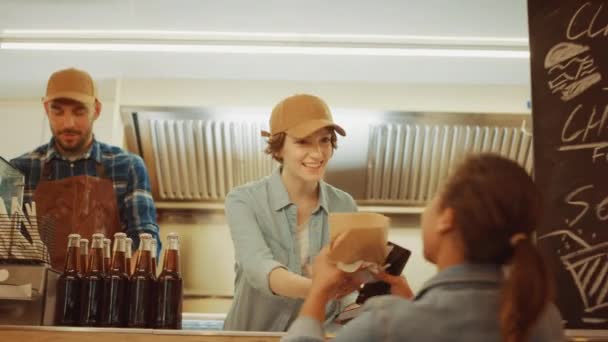 Food Truck Medewerker reikt een vers gemaakte hamburger uit aan een vrolijke jonge vrouw. Jongedame gebruikt NFC Mobile Payment om te betalen voor voedsel. Street Food Truck verkoopt hamburgers in een moderne hippe buurt. — Stockvideo