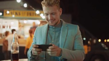 Yakışıklı Genç Adam Günlük Takımla Çok Bağımlı Online Video Oyunu Oynuyor. Bir Outdoor Street Food Cafe 'de bir masada otururken akıllı telefon kullanıyor. İndirdiği Uygulama.