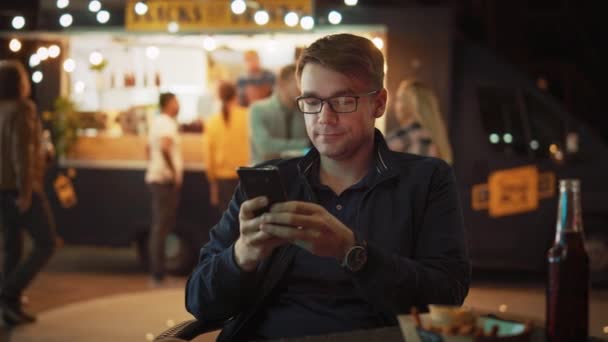 Hombre joven guapo en gafas está utilizando un teléfono inteligente mientras está sentado en una mesa en un café de comida al aire libre calle y comer papas fritas. Está navegando por Internet o las redes sociales, publicando una actualización de estado . — Vídeo de stock