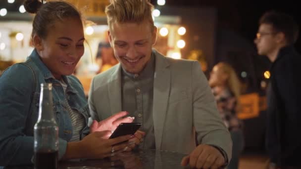 Mooi jong stel is het gebruik van een smartphone tijdens het zitten aan een tafel in een Outdoors Street Food Cafe. Ze surfen op internet of sociale media, kijken video 's. Ze zijn gelukkig en glimlachen.. — Stockvideo
