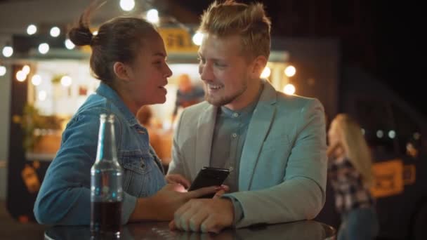 Hermosa pareja joven está utilizando un teléfono inteligente mientras se sienta en una mesa en un café de comida al aire libre de la calle. Están navegando por Internet o redes sociales, viendo videos. Son felices y sonríen . — Vídeos de Stock