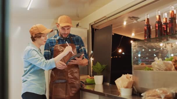 Udane Food Truck Mężczyzna i kobieta Pracownicy w brązowych czapkach są przy użyciu komputera typu tablet. Omawiają swoje zyski i plany biznesowe na przyszłość. Commercial Truck lub Kiosk Sprzedam Street Żywność i napoje. — Wideo stockowe