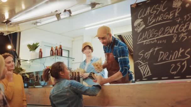 Empleado de camión de alimentos entrega una hamburguesa recién hecha a una joven y feliz hembra. Young Lady está pagando por comida con tarjeta de crédito sin contacto. Camión de comida callejera vendiendo hamburguesas en un vecindario moderno de la cadera — Vídeo de stock