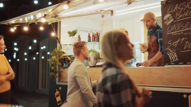 Food Truck Medewerker reikt een vers gemaakte hamburger uit aan een gelukkige jonge man. De man in een casual pak gebruikt NFC Mobile Payment Solution. Street Food Truck verkoopt hamburgers in een moderne hippe buurt. — Stockvideo