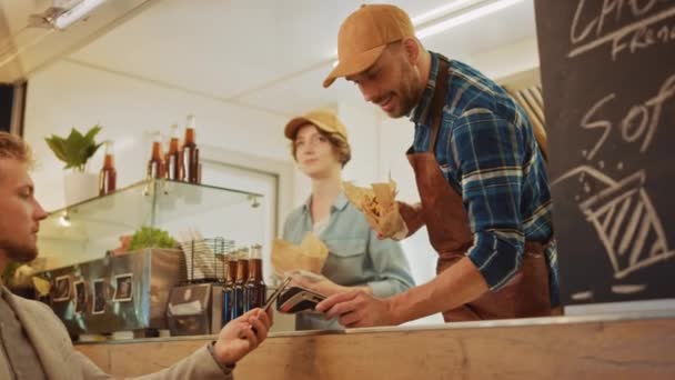 A Food Truck alkalmazottja frissen készített hamburgert ad egy boldog fiatal férfinak. Az alkalmi öltönyös férfi az NFC Mobil Fizetési Megoldást használja. Street Food Truck Burgereket árul egy modern csípős környéken. — Stock videók