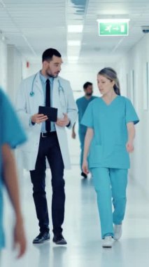 Cerrah ve kadın doktor hastane koridorunda yürürken hastanın sağlığı hakkında konuşurken dijital tablet bilgisayara danışıyorlar. Dikey Ekran Yönelimli Video Görüntüsü 9: 16