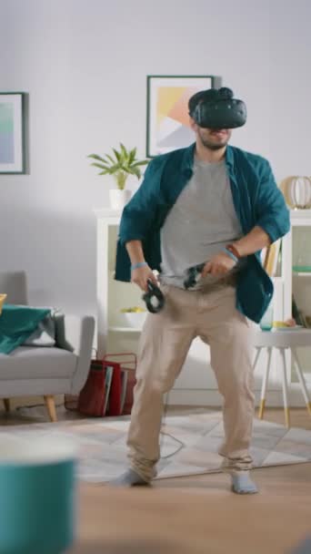 가상 현실 헤드셋을 착용하고 컨트롤러를 들고 활기찬 젊은이 가 집에서 비디오 게임을 하고 있다. 거실에서 VR 테니스를 연주하고 있습니다. 수직 화면 방향의 비디오 영상 — 비디오