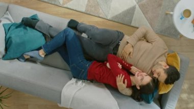 Genç ve güzel bir çift kanepeye uzanıyor. Adam Nazikçe Kızı Omuzundan Kucaklıyor. Yukarıya Bakar ve Gülümserler. Üst Manzara. Dikey Ekran Yönelimli Video Görüntüsü. 9: 16