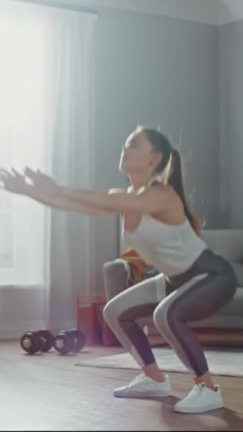 Forte et belle femme de fitness athlétique en vêtements de sport fait des exercices de squat dans son salon lumineux et spacieux avec intérieur minimaliste. Vidéo avec orientation verticale de l'écran — Video