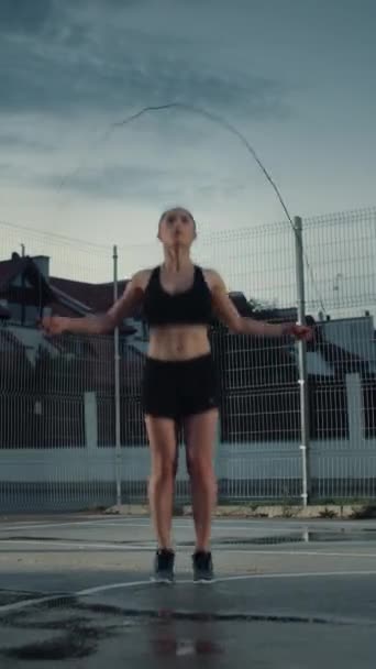 아름다운 체력의 소녀 줄넘기 / 점프 줄. 그녀는 울타리가 쳐진 야외 농구 코트에서 운동을 하고 있어요. 오후에 찍은 비가 내리고 — 비디오