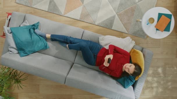 Young Girl in Red Jumper and Blue Jeans is Lying Down on a Sofa, Using a Smartphone. She is Happy and Smiles. Top View. Vertical Screen Orientation Video 9:16 — Stock Video