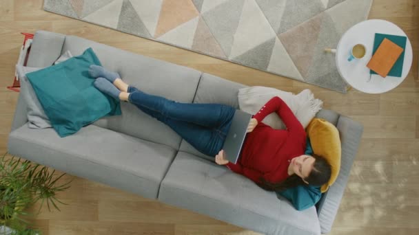 Young Girl in Red Blouse and Blue Jeans está acostado en un sofá, usando un ordenador portátil. Ella mira arriba y sonríe. Vista superior con zoom. Video con orientación vertical de la pantalla 9: 16 — Vídeo de stock