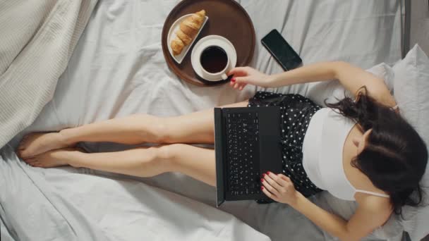 Young Millennial Girl Sentado en una cama por la mañana, utiliza computadora portátil y come cruasanes, bebe café para el desayuno. Zoom de arriba hacia abajo. Video con orientación vertical de la pantalla 9: 16 — Vídeos de Stock