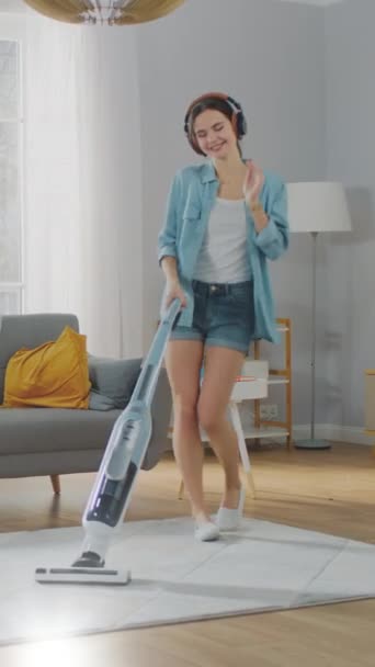 Young Beautiful Woman in Jeans Shirt and Shorts está escuchando música en sus cabeceras, bailando y limpiando al vacío una alfombra en una acogedora habitación en casa. Video con orientación vertical de la pantalla 9: 16 — Vídeo de stock