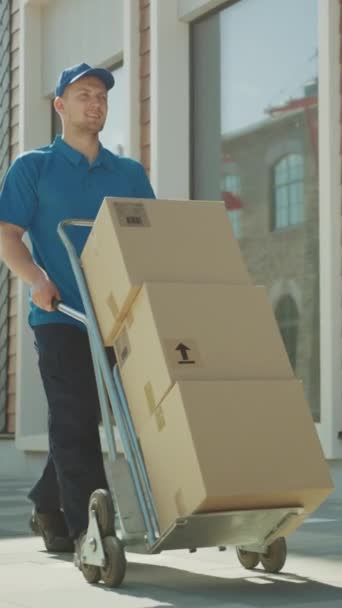 Happy Young Delivery Man empurra carrinho de mão caminhão cheio de caixas de papelão e pacotes para entrega. Correio profissional trabalhando de forma eficiente e rápida. Vídeo de Orientação de Tela Vertical 9: 16 — Vídeo de Stock