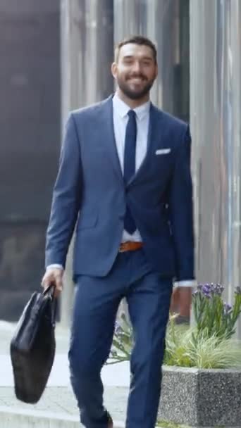 Hombre de negocios sonriente con una bolsa camina en un distrito de negocios. El tiempo es soleado Se ve guapo con su traje a medida. Vídeo de orientación vertical de la pantalla 9: 16 — Vídeos de Stock