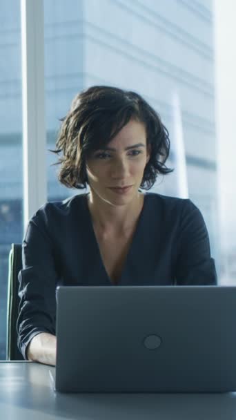 Empresaria confiada que trabaja en una computadora portátil en su oficina moderna. Elegante hermosa mujer haciendo trabajo importante. En la ventana Big City Business District View. Vídeo de orientación vertical de la pantalla 9: 16 — Vídeos de Stock