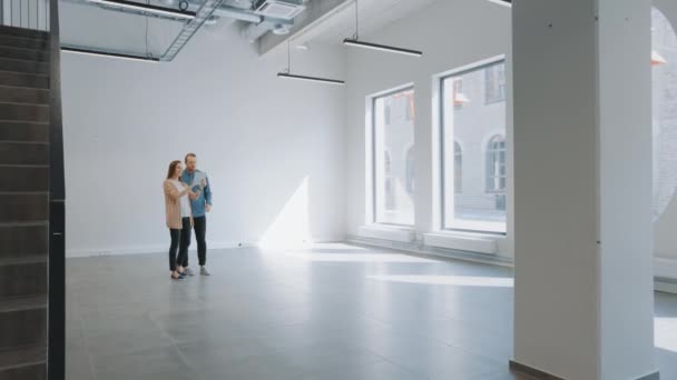 Young Hipster Man and Female Stand in an Empty White Office and Map it with an Augmented Reality Software on a Tablet. A luz solar brilha através de grandes janelas. Sala tem pontos de rastreamento para software de vídeo . — Vídeo de Stock