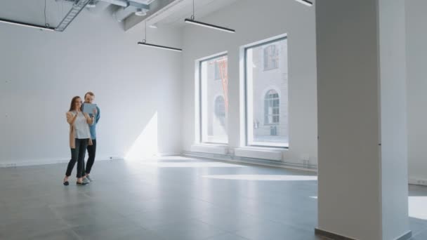 Młody Hipster Man i kobieta stoją w pustym, białym biurze i mapują go za pomocą rozszerzonego oprogramowania rzeczywistości na tablecie. Światło słoneczne świeci przez duże okna. Pokój posiada punkty śledzenia dla oprogramowania wideo. — Wideo stockowe