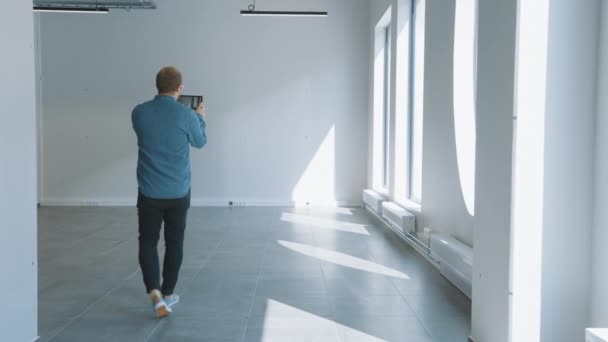 Mladý Hipster Man v džínové košili stojící v prázdné kanceláři a mapovat ji s rozšířenou realitou software na tabletu. Sluneční světlo svítí velkými okny. Místnost má sledovací body pro video software. — Stock video