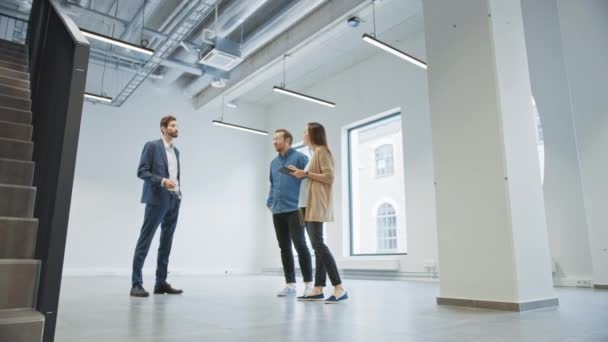 Agent immobilier montrant un nouvel espace de bureau vide aux jeunes hipsters masculins et féminins. Les entrepreneurs rencontrent le courtier avec une tablette et discutent de la facilité qu'ils souhaitent acheter ou louer. — Video