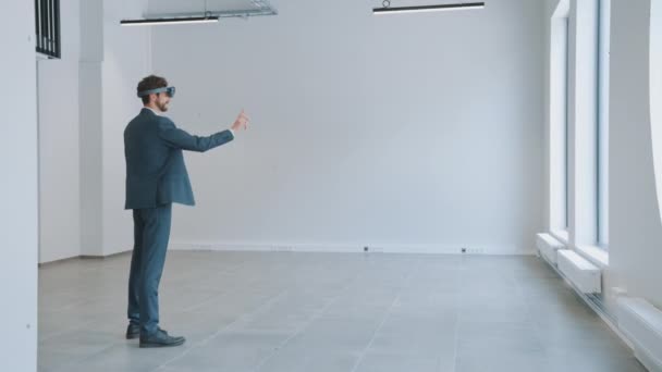 Empresario en gafas holográficas de realidad aumentada de pie en la oficina vacía y mapearlo. Empresario moviendo piezas virtuales alrededor del espacio. Sala tiene puntos de seguimiento para el software de vídeo . — Vídeos de Stock