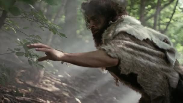 El cavernícola primitivo que lleva piel de animal sostiene la lanza de punta de piedra mira a su alrededor, explora el bosque prehistórico en una búsqueda de presas de animales. Neanderthal va a cazar en la selva — Vídeo de stock