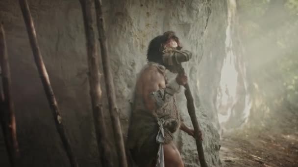 Oerholbewoner het dragen van een dierlijke huid houdt stenen getipt hamer komt uit de grot en kijkt rond prehistorische bos, klaar om te jagen dierlijke prooi. Neanderthal gaat jagen in de jungle. Arc shot — Stockvideo