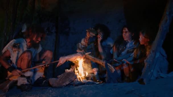 Neandertalare eller Homo Sapiens Familj Matlagning Animal Meat över Bonfire och sedan äta det. Stam av förhistoriska Hunter-samlare bär djurskinn Grillning och äta kött i grottan på natten — Stockvideo