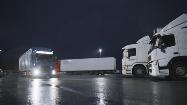 Blue Semi-Truck with Cargo Trailer Drives Off From Overnight Parking Space where Other Trucks are Standing. Long Haul Truck opustí parkoviště, Přeprava nákladu / zboží přes kontinent. Deštivá noc