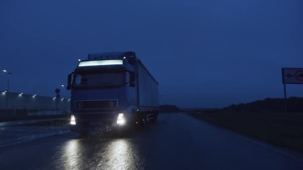 Long Haul Semi-Truck with Cargo Trailer Full of Goods Travels At Night on the Freeway Road, Driving Across Continent Through Rain, Fog, Snow. Industrial Warehouses Area. Front Following Shot