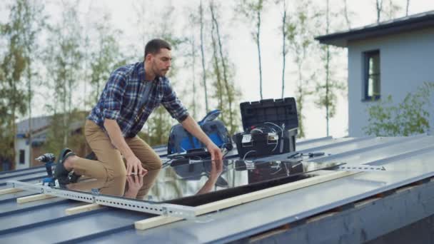 Hombre caucásico en camisa a cuadros está instalando paneles solares reflectantes negros a una base de metal con un taladro. Trabaja en el tejado de una casa en un día soleado. Concepto de Energía Renovable Ecológica en el Hogar. — Vídeos de Stock