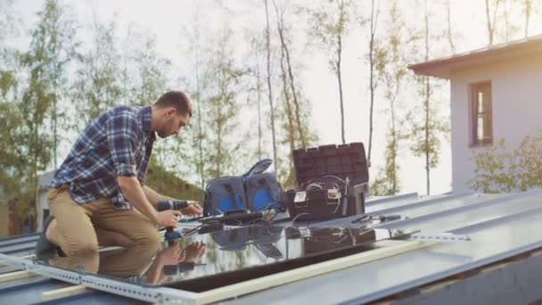 L'homme caucasien en chemise à carreaux installe des panneaux solaires réfléchissants noirs sur une base métallique avec une perceuse. Il travaille sur un toit de maison par une journée ensoleillée. Concept d'énergie renouvelable écologique à la maison. — Video