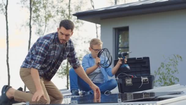 Far och son Installera solpaneler till en metallbas med en borrmaskin. De arbetar på ett hus tak på en solig dag och High Five. Begreppet ekologisk förnybar energi i hemmet och kvalitet familjetid. — Stockvideo