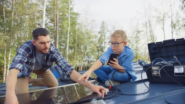 Père et Fils Installation de panneaux solaires sur une base métallique. Ils travaillent avec le câblage sur un toit de maison par une journée ensoleillée. Boy regarde un manuel sur un smartphone. Père relie les fiches et ils High Five . — Video