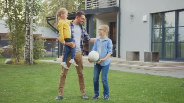 Retrato de una familia feliz de tres: padre, hija, hijo. Están posando frente a la cámara en un césped al lado de su casa de campo. Papá sostiene a la chica en sus brazos. Chico está sosteniendo un fútbol. — Vídeos de Stock