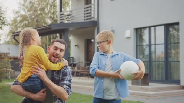 Porträtt av en lycklig familj på tre: far, dotter, son. De sätter sig framför kameran på en gräsmatta bredvid deras hus. Pappa håller flickan i sina armar. Boy håller i en fotboll. — Stockvideo