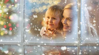Noel arifesinde Anne ve Kızı Karlı Efekt ile Pencereden Bakıyor. Pencerede Çiçek Parlıyor.