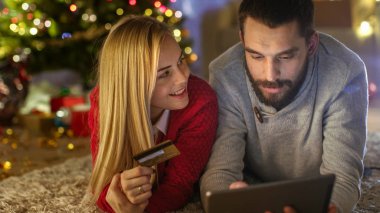 Mutlu çift yalan Noel ağacı, kadın tutar kredi kartı ve adam altında halı üzerinde satın hediyelerini Tablet bilgisayarda.