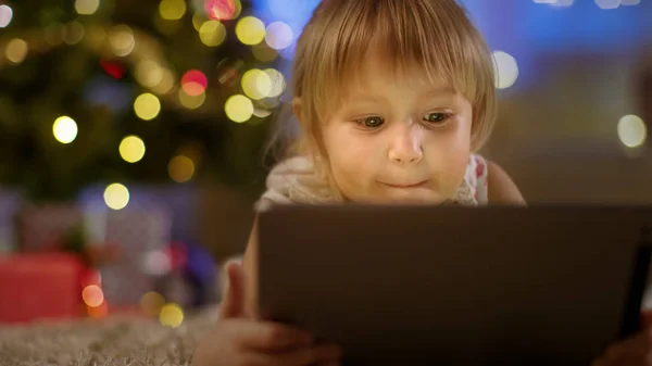 Schattig klein meisje met tabletcomputer in haar handen ligt op het tapijt onder de kerstboom. — Stockfoto
