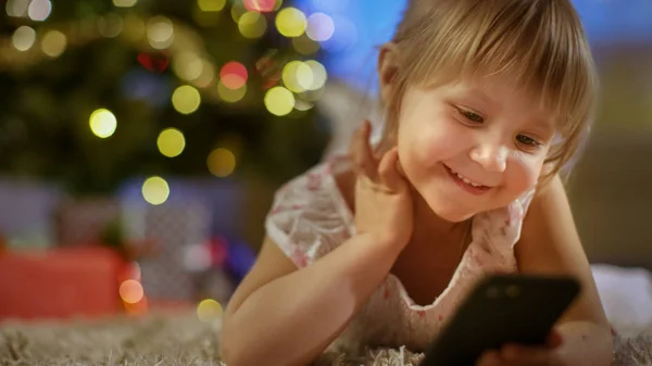 Schattig klein meisje met Smartphone in haar handen ligt op het tapijt onder de kerstboom. — Stockfoto