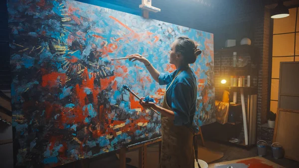 Talentierte Künstlerin arbeitet an abstrakter Ölmalerei, mit Pinsel schafft sie ein modernes Meisterwerk. dunkles und chaotisches Kreativstudio, in dem große Leinwand auf Staffelei beleuchtet steht. — Stockfoto