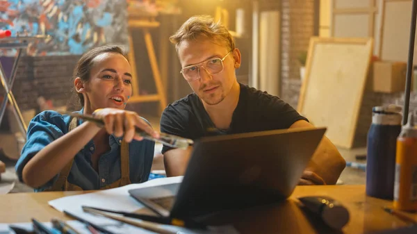 Paar artiesten zitten in hun studio gebruik laptop computer, werken aan een project, praten en glimlachen. Schilder en haar partner doen onderzoek. Authentieke workshop met olieverfschilderijen en gereedschap — Stockfoto