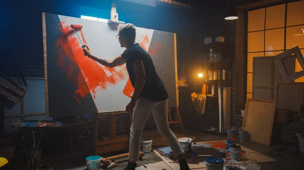 Talented Male Artist Working on an Abstract Painting, Uses Industrial Roller To Paint Daringly Emotional Modern Picture. Dark Creative Studio Large Canvas Stands on Easel Illuminated. — ストック写真
